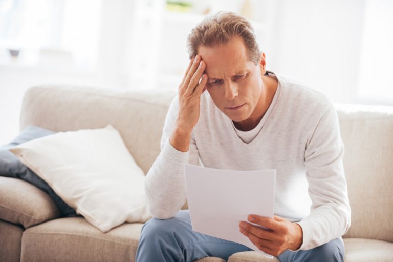 Bad news. Depressed mature man holding paper and looking at it while sitting on the couch at home